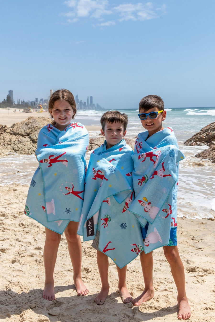 Three children wrapped in towels on a beach with a city skyline in the background. The Elf On The Shelf Gift Idea
