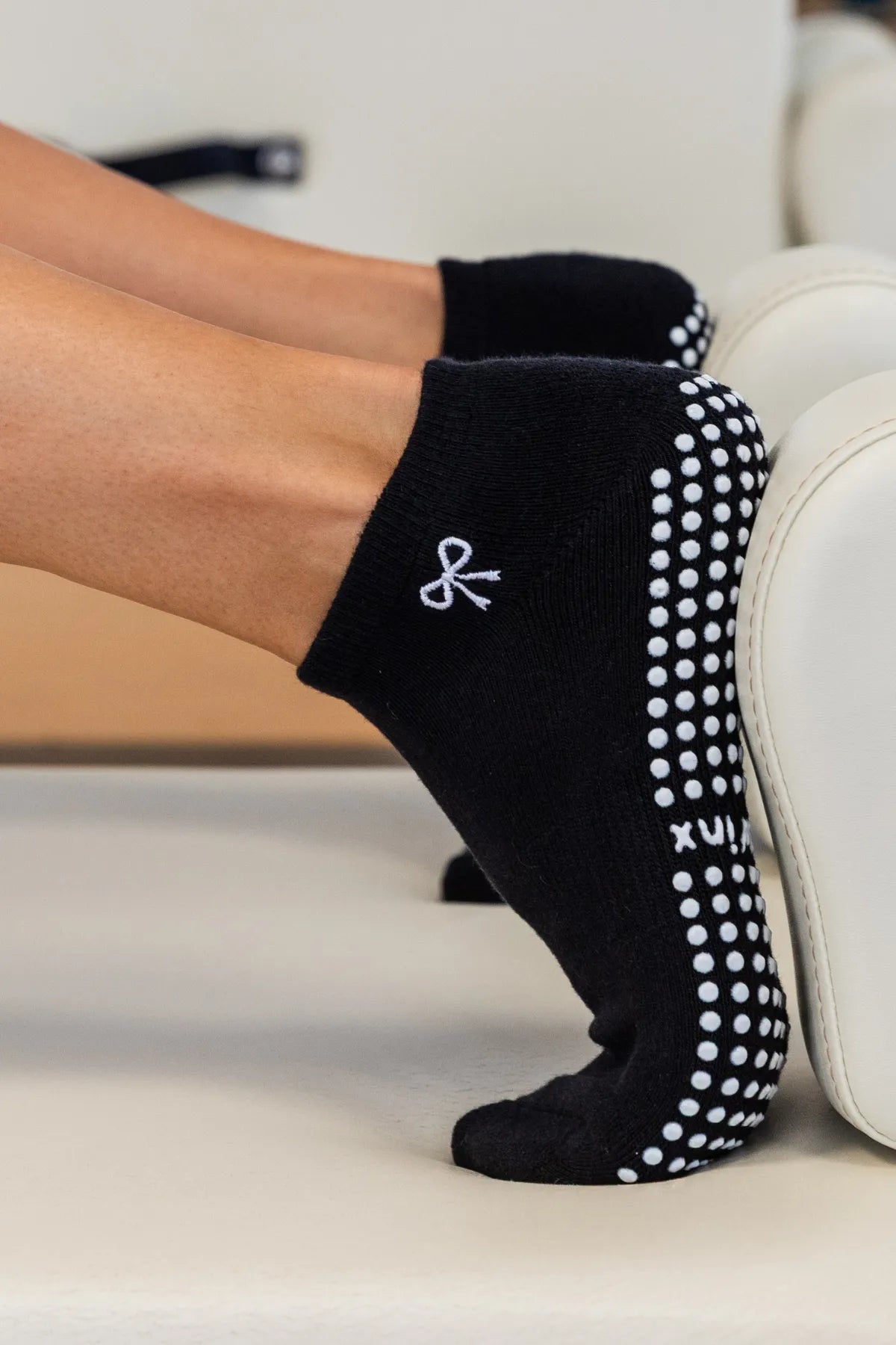 Black ankle grip socks with white grip dots and a white bow logo, worn on a person's feet resting on a reformer. cheeky Winx non slip socks for Pilates 